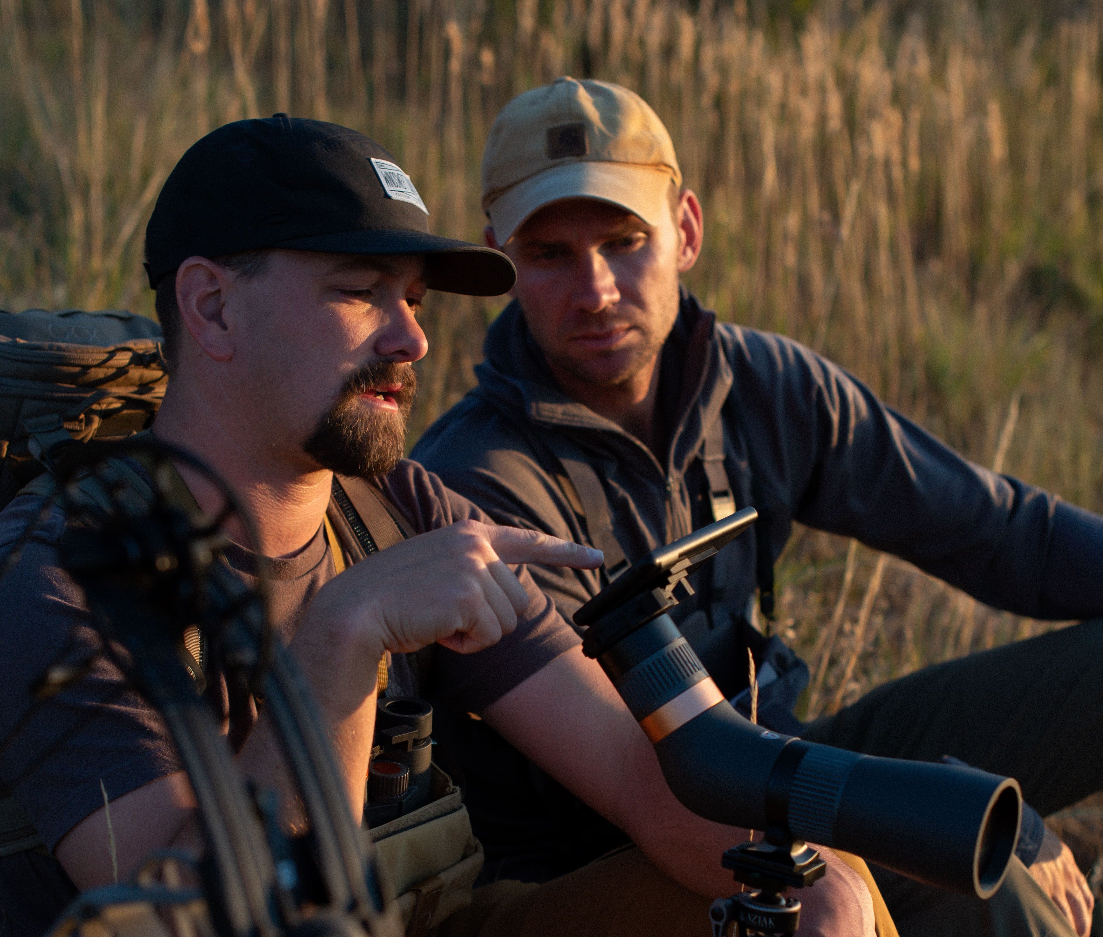 Two men in a field with one using a bow and arrow, the other with binoculars.